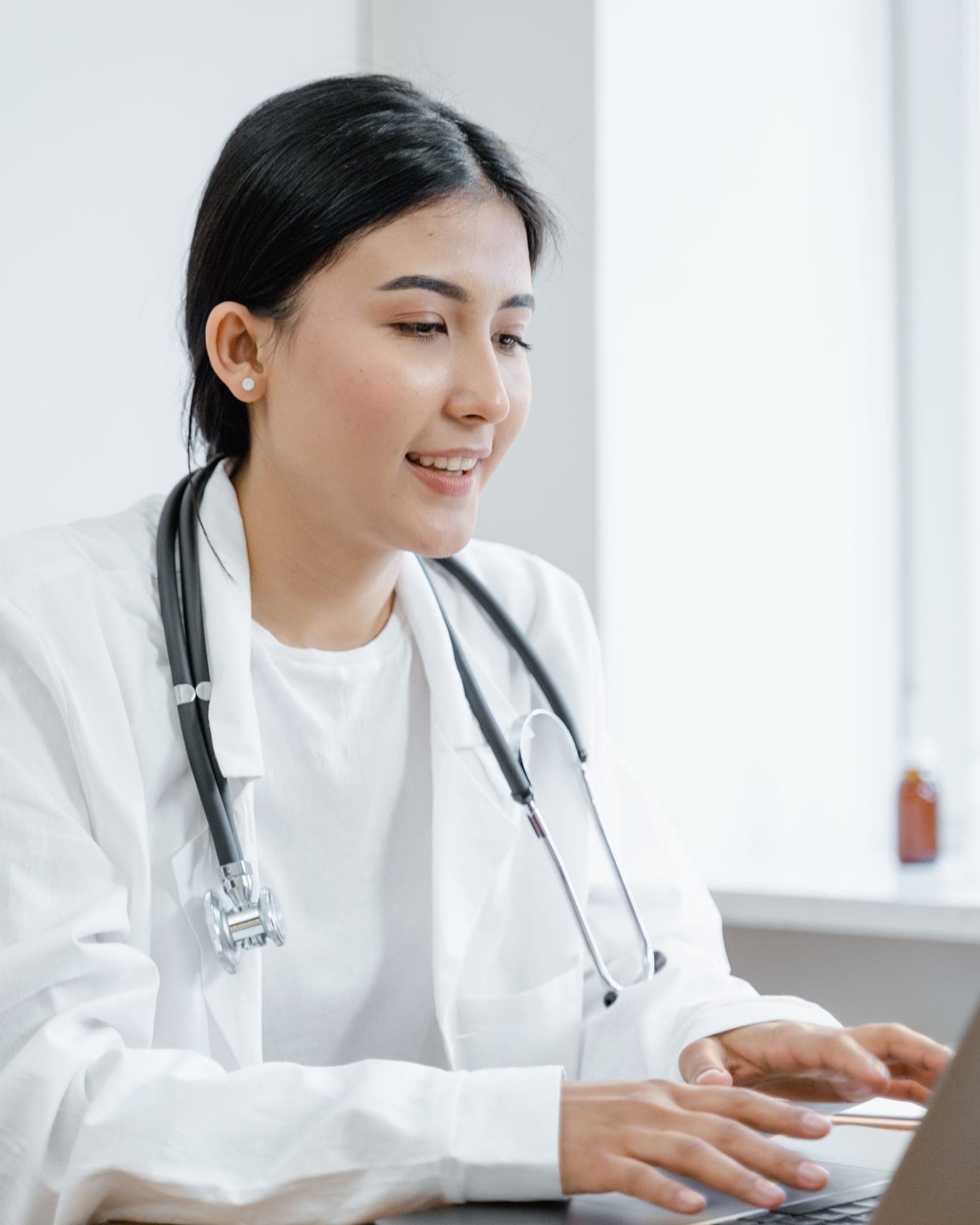 A doctor wearing a white coat and stethoscope, working at a laptop.